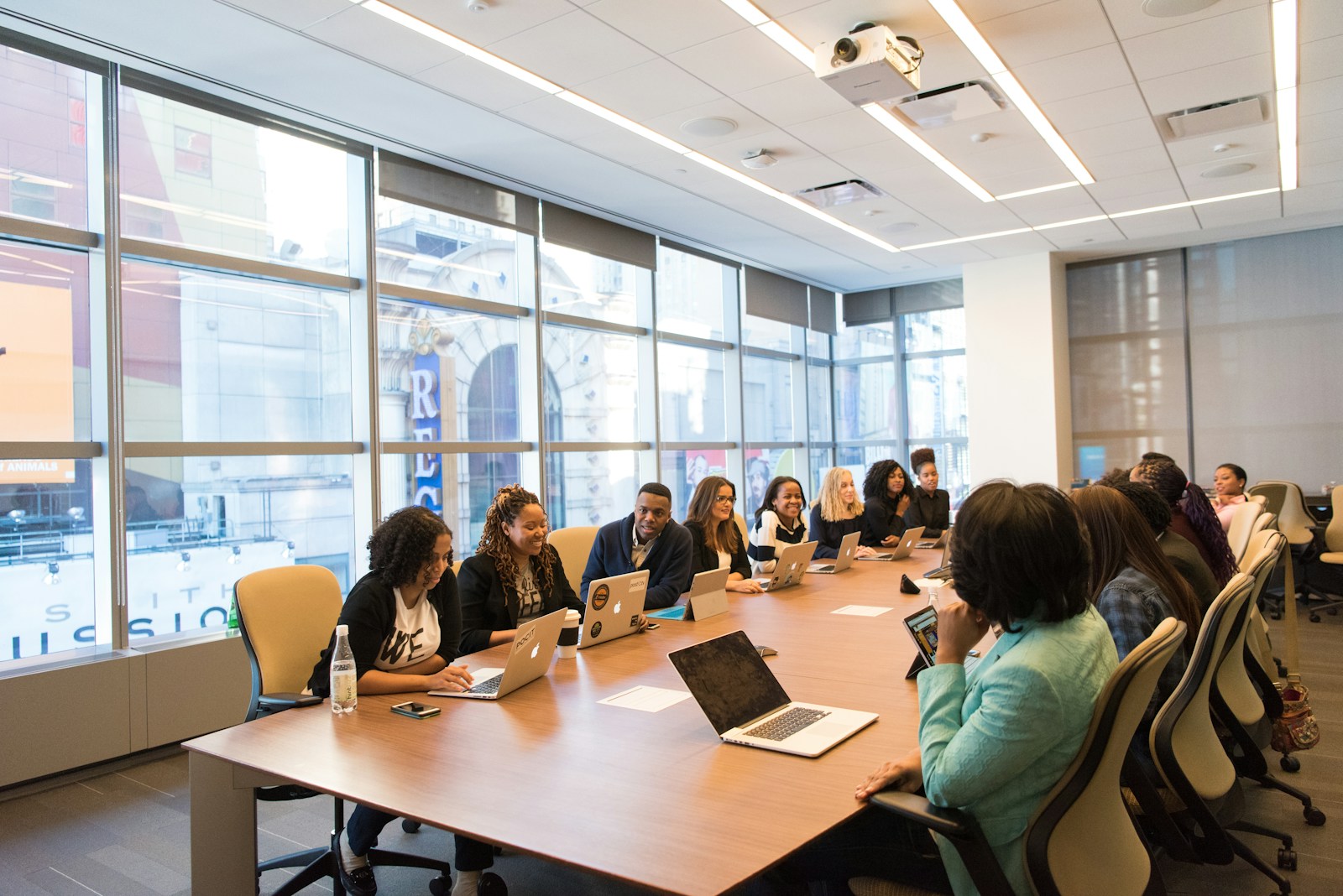 group of people sitting at a rectangular wooden table with laptops in a conference room with large windows